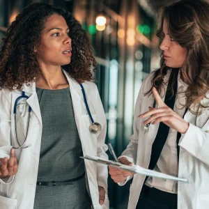 Two female doctors in white lab coats discussing patient care in a hospital hallway, representing teamwork challenges in healthcare environments.